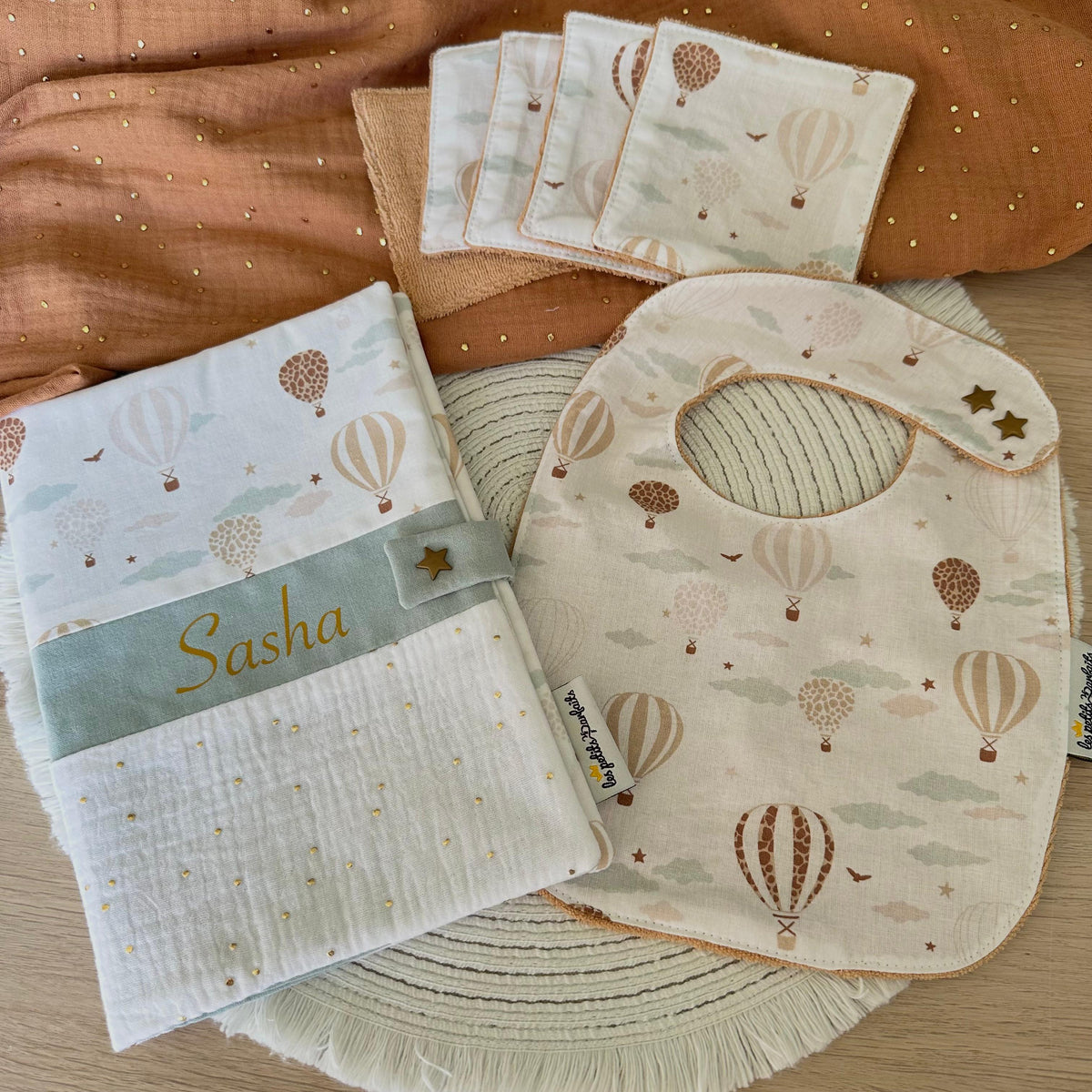 Coffret naissance bébé univers montgolfières composé d'un protège carnet de santé personnalisé avec prénom (police et coloris au choix), d'un bavoir bébé évolutif double pression étoile en éponge bio et de 5 lingettes lavables zéro déchet.